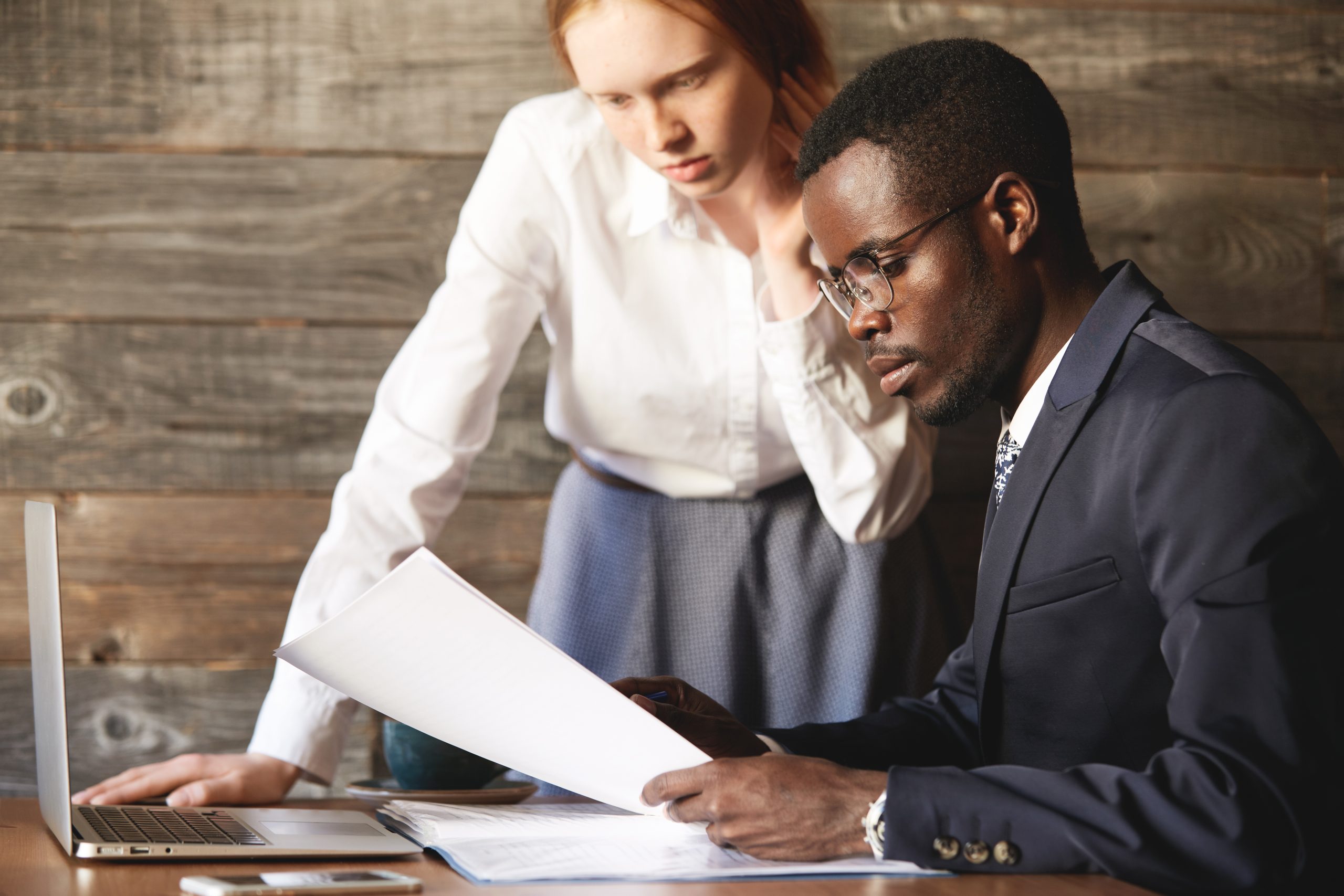 Portrait of African American businessman wearing formal suit and glasses, sitting at the wooden table at a cafe, looking at documents, checking a report, made by his young redhead Caucasian secretary